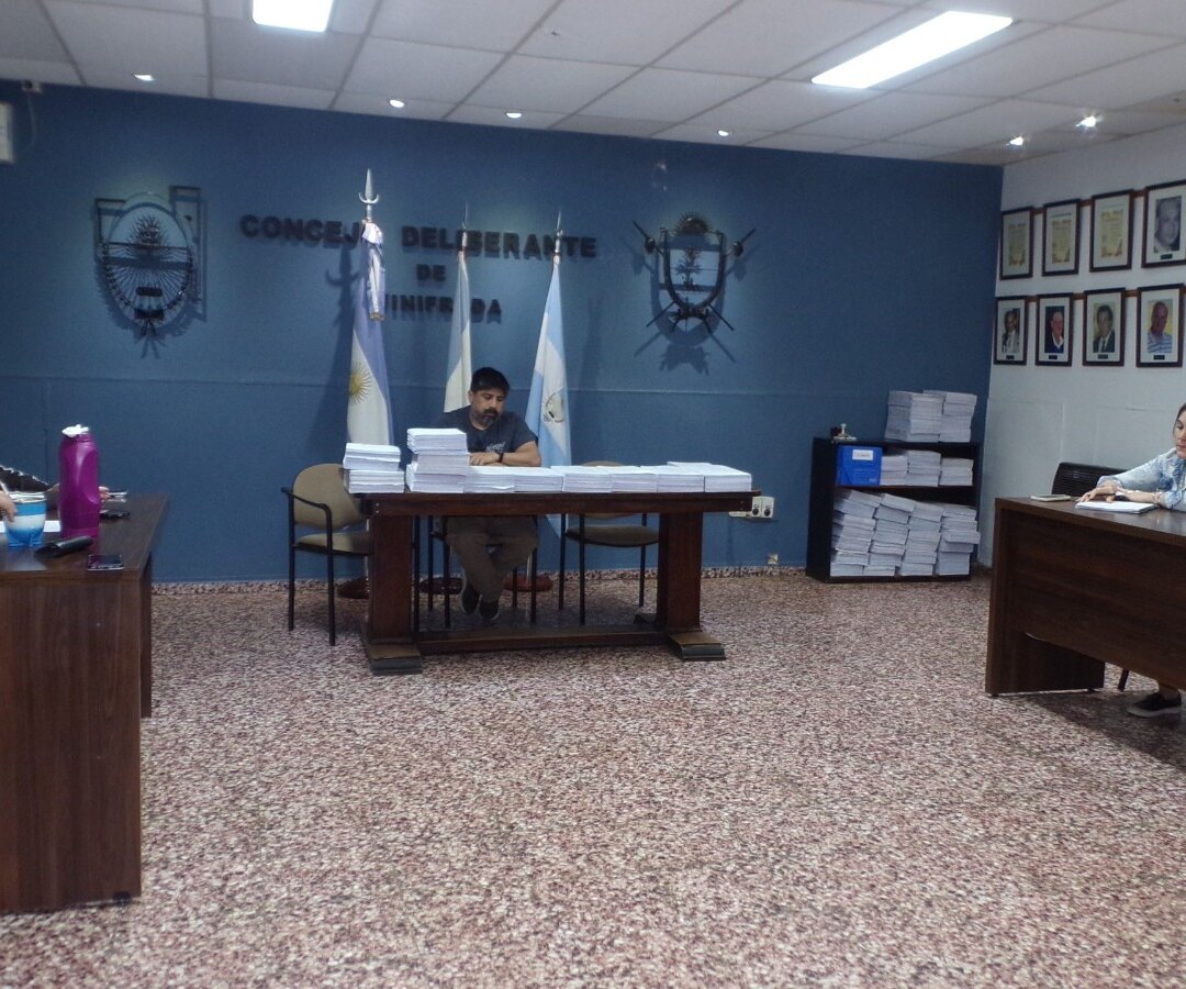 Sesión en el recinto del Concejo Deliberante de Winifreda, con concejales en sus bancas y pilas de documentación sobre la mesa central