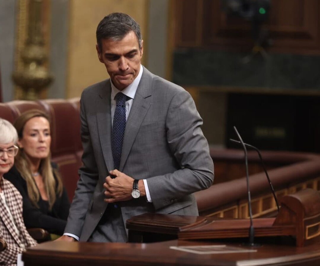 Pedro Sánchez de pie frente al atril del Congreso de los Diputados, con dos legisladoras sentadas detrás