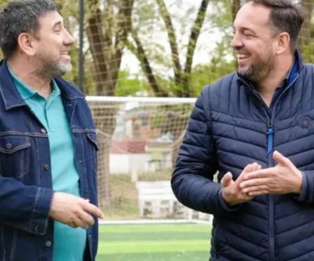 Dos hombres conversan y sonríen al aire libre, sobre una cancha de césped sintético con un arco de fútbol detrás