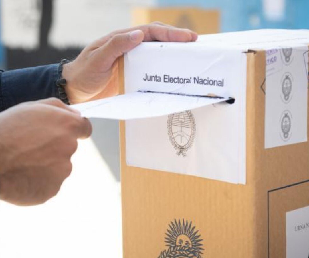 Manos introducen un sobre en una urna de cartón con la faja de la Junta Electoral Nacional
