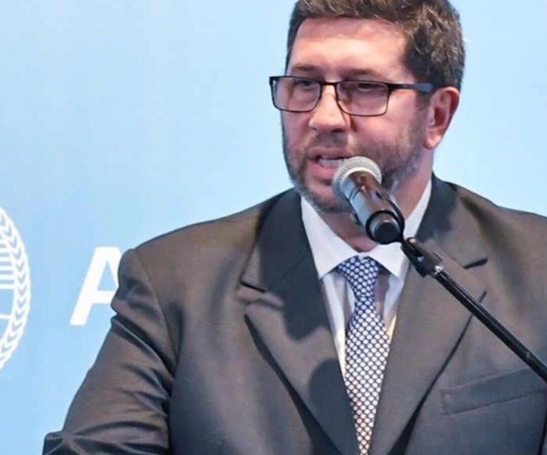 Hombre de traje y anteojos habla ante un micrófono frente a un panel celeste con el escudo nacional