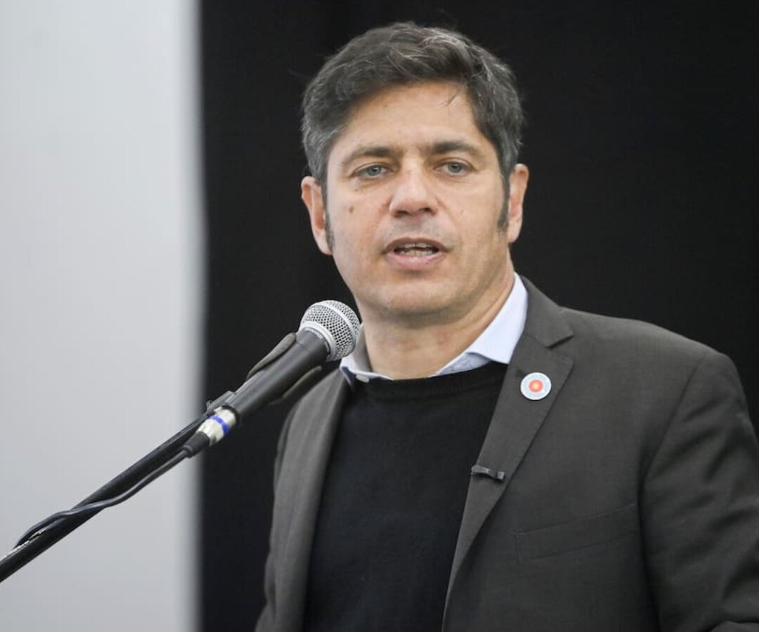 Hombre de saco gris y pulóver oscuro habla ante un micrófono durante un acto, sobre un fondo oscuro